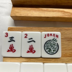 May include: Three white tiles with black and red Chinese characters and a black and green design. The tiles are labeled "3", "2", and "JOKER". The tiles are sitting on a wooden rack.