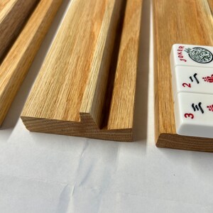 May include: Wooden rack with four slots for holding mahjong tiles. The rack is made of light brown wood and has a smooth finish. Three tiles are visible in the rack, showing the numbers "2", "3", and "Joker".