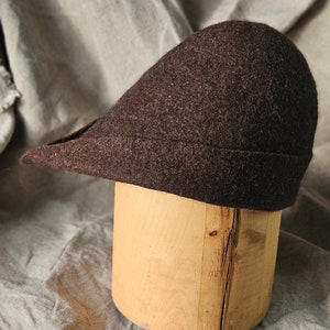 May include: A dark brown wool hat with a curved brim, displayed on a light brown wooden hat block. The hat has a rounded crown and a slightly angled brim. The background is a textured, neutral-colored fabric.