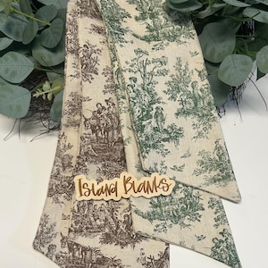 Puede incluir: Tres diademas de tela con un estampado toile de Jouy en marrón y verde. Las diademas están dobladas y dispuestas en forma de abanico. Un letrero de madera que dice "Island Blanks" está en el centro.