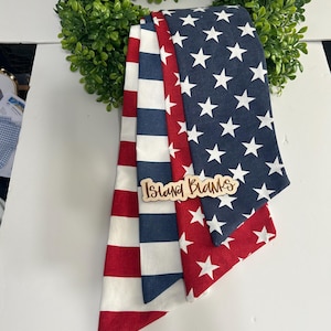 May include: A stack of three patriotic-themed fabric items, featuring red and white stripes, blue with white stars, and red with white stars. A wooden sign with the text "Island Blanks" is placed on top. The items are set against a green wreath.