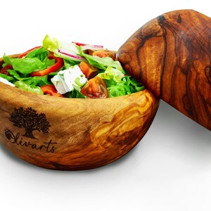 Peut inclure: Un saladier en bois avec un couvercle, tous deux en bois d'olivier. Le saladier est rempli d'une salade de laitue, tomates, feta et oignon rouge. Le saladier porte un logo représentant un olivier avec le texte "Olivarts" en dessous.