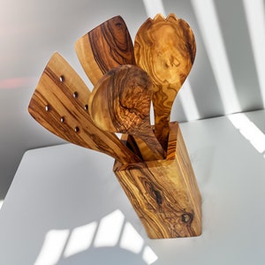 Peut inclure: Un ensemble de quatre ustensiles de cuisine en bois aux motifs de grain distincts, comprenant une spatule ajourée, une spatule pleine, une cuillère ronde et une fourchette à salade, rangés dans un porte-ustensiles rectangulaire assorti.