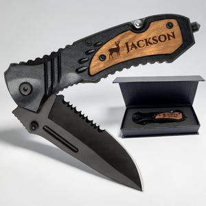 Peut inclure: Un couteau pliant avec une lame dentelée noire mate et un manche noir doté d'une incrustation en bois gravée d'une silhouette de cerf et du nom JACKSON. Un couteau assorti est présenté dans une boîte cadeau rectangulaire noire ouverte.