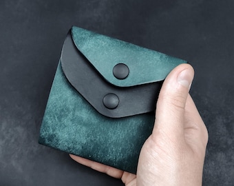 May include: A teal and black leather wallet with a snap closure. The wallet is held in a hand against a dark background. The teal leather has a textured appearance.