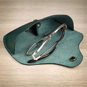 Könnte beinhalten: Ein türkisfarbenes Lederetui für Brillen mit einer silberfarbenen Metallgestellbrille im Inneren. Das Etui hat eine Klappe, die mit einem Druckknopf geschlossen wird.