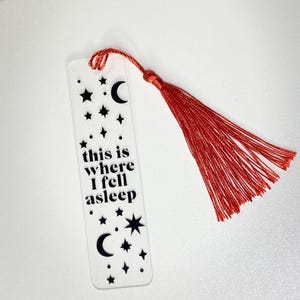 Puede incluir: Marcapáginas de acrílico transparente con borla roja. El marcapáginas presenta un diseño de estrellas, lunas y la frase "this is where I fell asleep" en letras negras. Un artículo decorativo para los amantes de los libros.