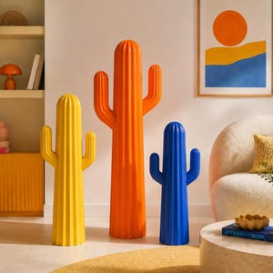 Op de afbeelding: Drie staande cactussculpturen in heldere, effen kleuren. De linker cactus is geel, de middelste is oranje en de rechter is blauw. Elk beeld heeft een geribbelde, verticale textuur en twee zijarmen die naar buiten steken.