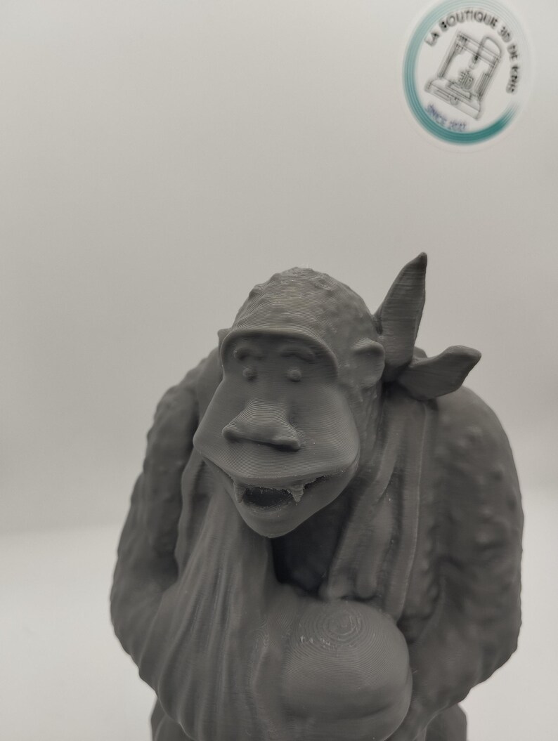 May include: A gray 3D printed figurine of a monkey wearing a bandana and holding a round object. The monkey has a wide mouth and large teeth.