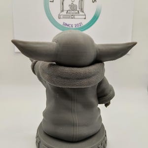 Puede incluir: Una figura impresa en 3D de color gris de un personaje de la franquicia de Star Wars, The Mandalorian. El personaje lleva una t&uacute;nica marr&oacute;n con capucha y est&aacute; de pie sobre una base con las palabras "This is the way" impresas en ella.