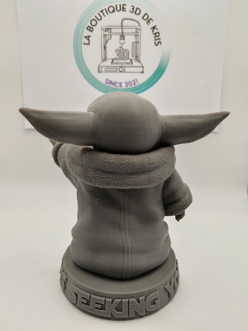Puede incluir: Una figura impresa en 3D de color gris de un personaje de la franquicia de Star Wars, The Mandalorian. El personaje lleva una t&uacute;nica marr&oacute;n con capucha y est&aacute; de pie sobre una base con las palabras "This is the way" impresas en ella.