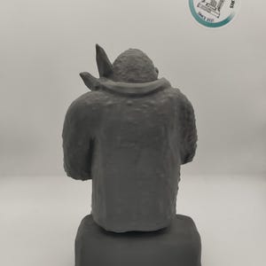 May include: A gray 3D printed figurine of a creature with a large head and a small body. The figurine is sitting on a gray base.