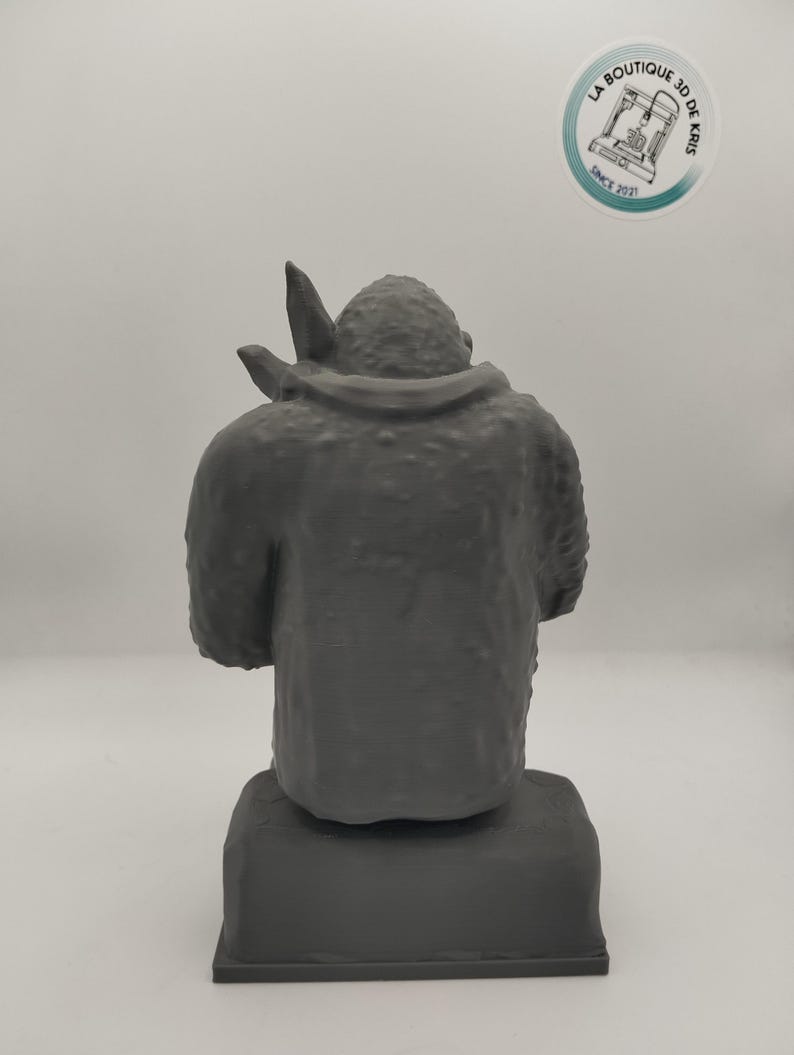 May include: A gray 3D printed figurine of a creature with a large head and a small body. The figurine is sitting on a gray base.