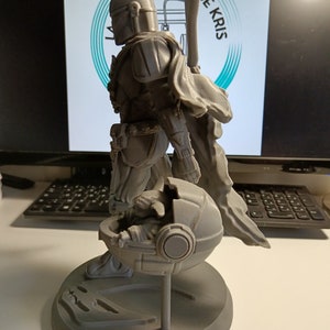 May include: A gray 3D printed figurine of a Star Wars character, The Mandalorian, holding a small, gray, round object. The Mandalorian is wearing a helmet and armor, and the object appears to be a pod or a container. The figurine is standing on a gray base.
