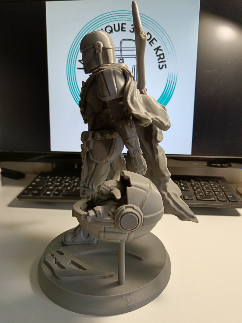 May include: A gray 3D printed figurine of a Star Wars character, The Mandalorian, holding a small, gray, round object. The Mandalorian is wearing a helmet and armor, and the object appears to be a pod or a container. The figurine is standing on a gray base.