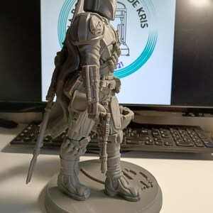 May include: A gray 3D printed figurine of a Mandalorian character from the Star Wars franchise. The character is wearing a helmet and armor, and is holding a weapon. The figurine is standing on a round base.