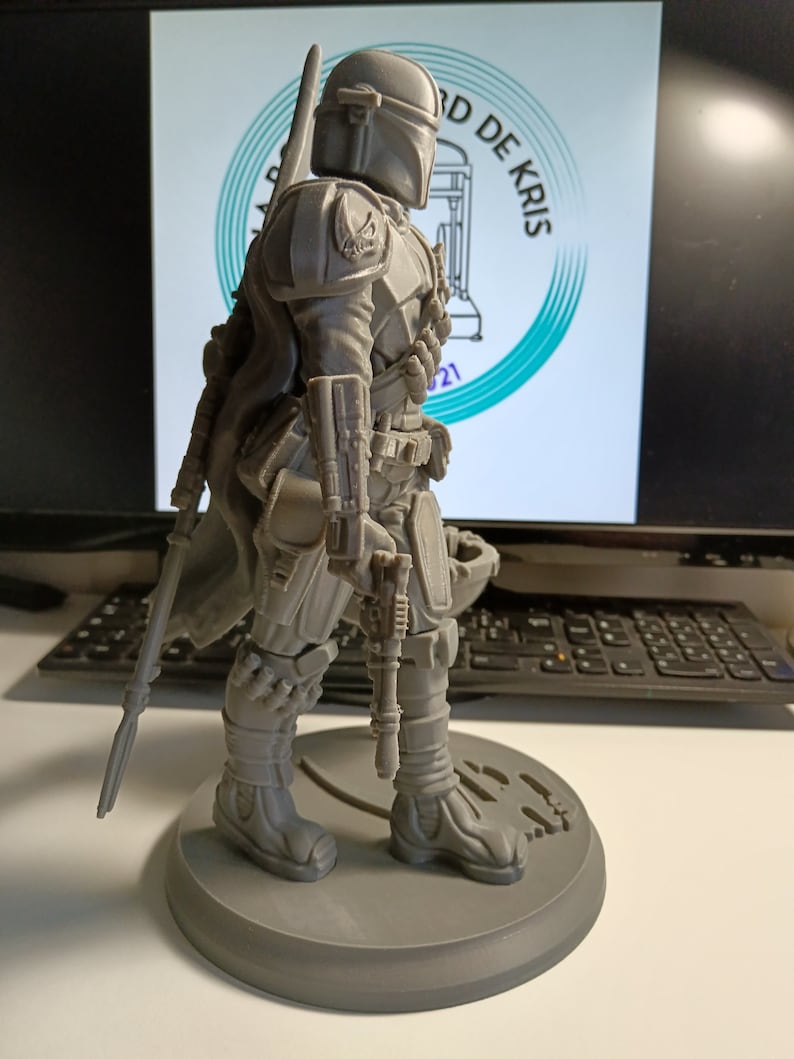 May include: A gray 3D printed figurine of a Mandalorian character from the Star Wars franchise. The character is wearing a helmet and armor, and is holding a weapon. The figurine is standing on a round base.
