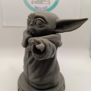 Puede incluir: Una figura impresa en 3D de color gris de un personaje de la franquicia Star Wars, conocido como "The Child" o "Baby Yoda". La figura est&aacute; de pie sobre una base redonda con las palabras "You Seek It" inscritas en ella.