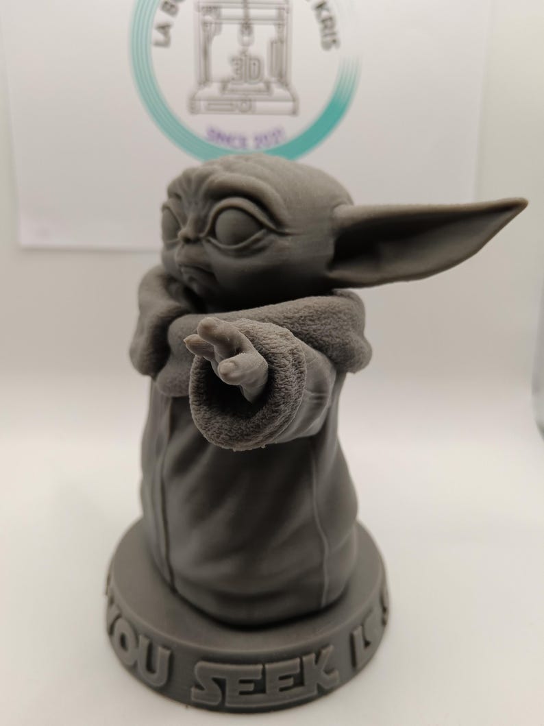 Puede incluir: Una figura impresa en 3D de color gris de un personaje de la franquicia Star Wars, conocido como "The Child" o "Baby Yoda". La figura est&aacute; de pie sobre una base redonda con las palabras "You Seek It" inscritas en ella.