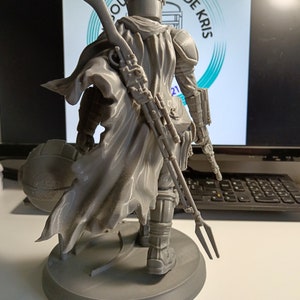 May include: A gray 3D printed figurine of a character from the Star Wars franchise, wearing a helmet and a cape, holding a weapon. The figurine is standing on a round base.