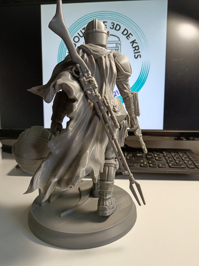 May include: A gray 3D printed figurine of a character from the Star Wars franchise, wearing a helmet and a cape, holding a weapon. The figurine is standing on a round base.