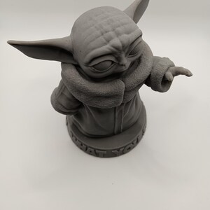 Puede incluir: Una figura impresa en 3D de color gris de un personaje de la franquicia Star Wars, conocido como "The Child" o "Baby Yoda". La figura lleva un abrigo marr&oacute;n y tiene una cabeza grande con orejas grandes. La base de la figura dice "What you'd do".