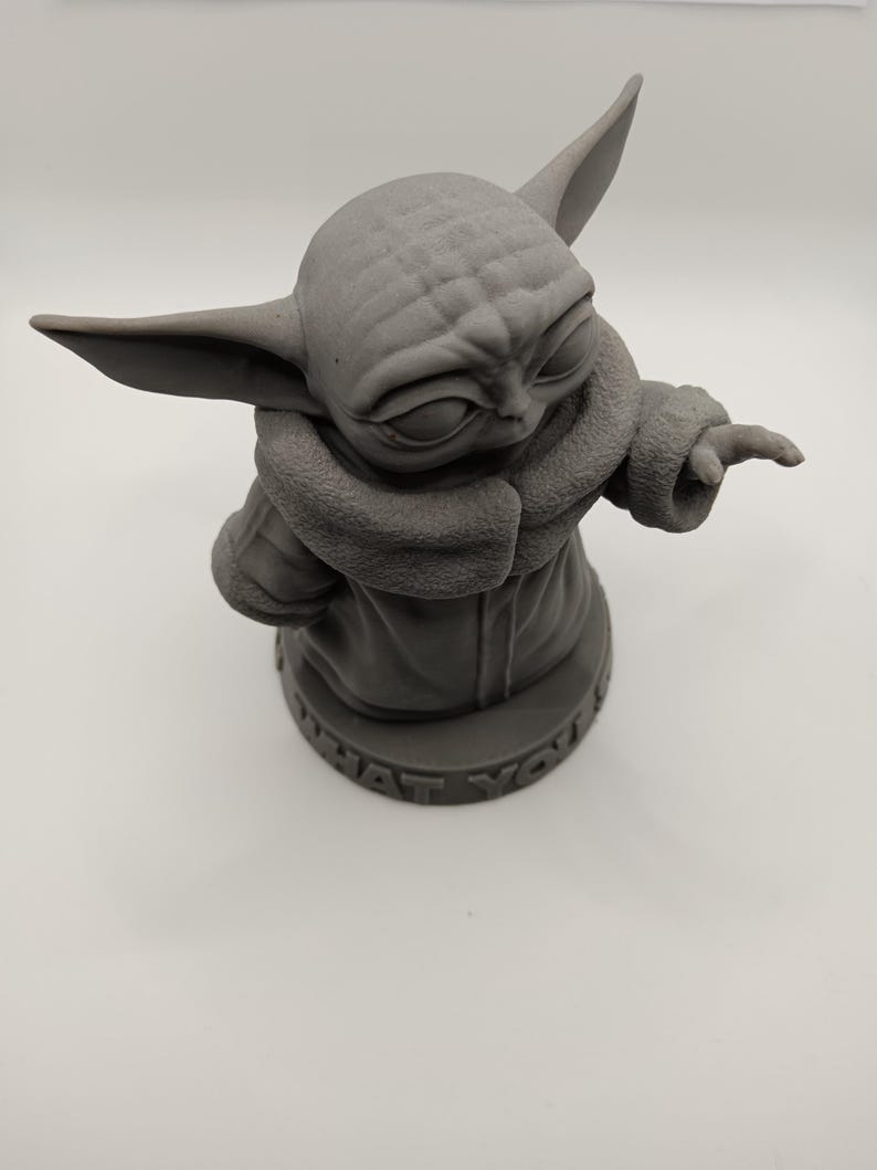 Puede incluir: Una figura impresa en 3D de color gris de un personaje de la franquicia Star Wars, conocido como "The Child" o "Baby Yoda". La figura lleva un abrigo marr&oacute;n y tiene una cabeza grande con orejas grandes. La base de la figura dice "What you'd do".