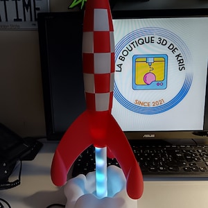 Könnte beinhalten: Eine rot-weiße raketenförmige Lampe mit einem weißen Wolkensockel. Die Lampe ist mit einem blau-weißen Licht beleuchtet. Die Lampe steht auf einem Schreibtisch mit einem Laptop und einem Telefon.