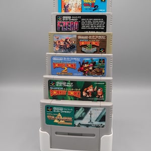 Könnte beinhalten: Ein vertikaler Stapel von sechs Super Famicom Videospielkassetten in einem weißen Kunststoffständer. Die Spiele umfassen Dragon Ball Z 2, F-Zero, Super Donkey Kong 3, Super Donkey Kong 2, Super Donkey Kong und einen Zelda-Titel, jeweils mit bunten Etiketten.