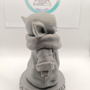 Puede incluir: Una figura impresa en 3D de color gris de un personaje de la franquicia Star Wars, conocido como "The Child" o "Baby Yoda". La figura lleva una t&uacute;nica marr&oacute;n y est&aacute; de pie sobre una base con el texto "I am you what" impreso en ella.
