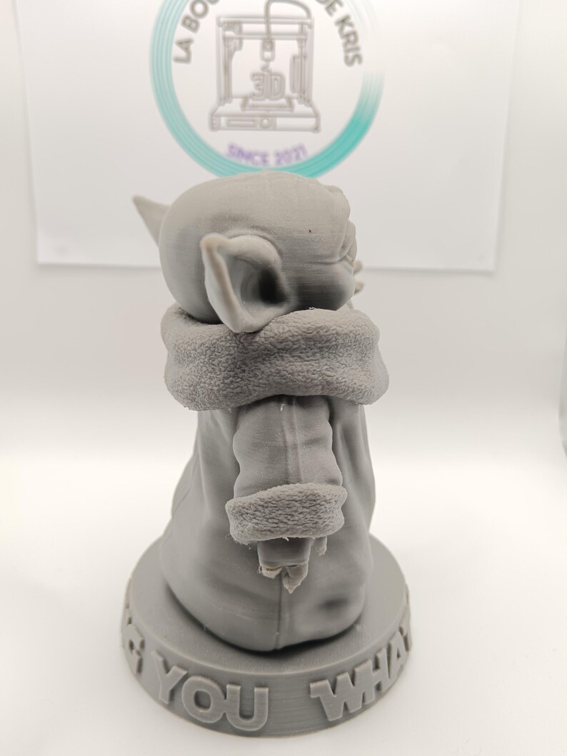 Puede incluir: Una figura impresa en 3D de color gris de un personaje de la franquicia Star Wars, conocido como "The Child" o "Baby Yoda". La figura lleva una t&uacute;nica marr&oacute;n y est&aacute; de pie sobre una base con el texto "I am you what" impreso en ella.
