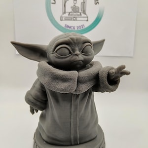 Puede incluir: Una figura impresa en 3D de color gris de un personaje de la franquicia Star Wars, conocido como "The Child" o "Baby Yoda". La figura est&aacute; de pie sobre una base con el texto "What You Say" impreso en ella.