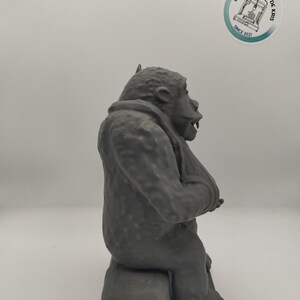 May include: A gray 3D printed figurine of a gorilla sitting on a small platform. The gorilla is in a seated position with its arms crossed and its head tilted slightly to the side.