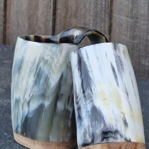 Peut inclure: Deux tasses &agrave; boire en corne avec des bases en bois. Les tasses sont faites de corne naturelle et pr&eacute;sentent un motif marbr&eacute; unique dans des tons de gris, de blanc et de noir.