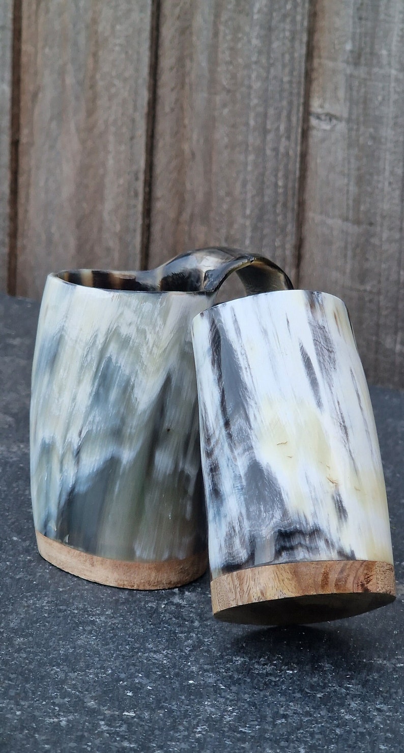 Peut inclure: Deux tasses &agrave; boire en corne avec des bases en bois. Les tasses sont faites de corne naturelle et pr&eacute;sentent un motif marbr&eacute; unique dans des tons de gris, de blanc et de noir.