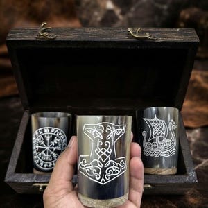 Puede incluir: Tres cuernos para beber con diseños grabados en blanco, incluyendo un Vegvisir, un martillo de Mjolnir y un barco vikingo, presentados en una caja de madera marrón oscuro con cierres metálicos. Los cuernos tienen tonos grises y negros.