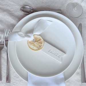 May include: A white ceramic place card with the name "Jamie" written on it. A dried lemon slice is attached to the place card with a white ribbon.