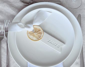 May include: A white ceramic place card with the name "Jamie" written on it. A dried lemon slice is attached to the place card with a white ribbon.