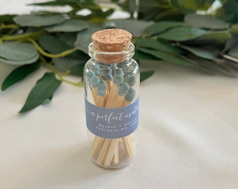 Può includere: Un piccolo barattolo di vetro pieno di fiammiferi di legno con punte blu. Il barattolo ha un tappo di sughero e un'etichetta che recita "a perfect match" con i nomi Rachel e Steve e la data 23 febbraio 2023.