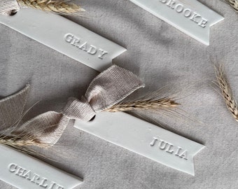 May include: Four white ceramic place card holders with a banner shape and a wheat stalk attached to each. The names "Grady", "Brooke", "Julia", and "Charlie" are embossed on the place cards.