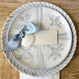May include: A decorative place setting featuring a white plate with a blue floral design and a matching smaller plate. A light blue ribbon, a seashell, and a name card with "AVA" are arranged on the plate. The plates are on a wooden surface.