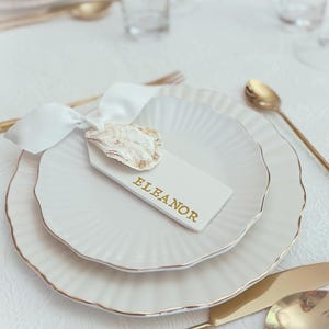 May include: White place setting with a gold rim, featuring a white plate with a gold rim, a white napkin with a white oyster shell tied with a white ribbon, and a white place card with the name "Eleanor" written in gold lettering.
