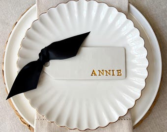 May include: A white scalloped-edge plate with a gold rim, holding a white tag with the name "ANNIE" in gold lettering. A black satin ribbon is tied around the tag. The plate rests on a woven charger and linen napkin.