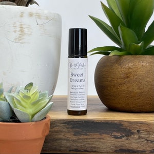 May include: A brown glass roller bottle with a white label that reads "Wanita Milen Naturally - Sweet Dreams - A drop or two to help you sleep - Essential Oils: Bergamot, Chamomile, Cedarwood, Clary Sage, Eucalyptus, Frankincense, Lavender, Mandarin Orange Sweet, Vetiver, Ylang Ylang - Other Ingredients: Olive Oil"