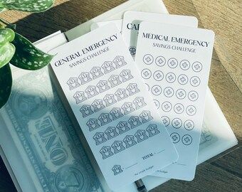 Pode incluir: Cartões brancos de desafio de poupança com texto e ilustrações pretas. Um cartão diz "GENERAL EMERGENCY SAVINGS CHALLENGE" com ilustrações de bancos. Outro diz "MEDICAL EMERGENCY SAVINGS CHALLENGE" com sinais de mais. Uma nota de um dólar é visível.