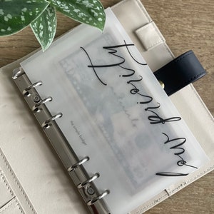 May include: A cream-colored binder with a black strap and gold button closure. A clear plastic envelope inside the binder has the words "Trust and Save" in black script. The text "the simple budget" is also visible.