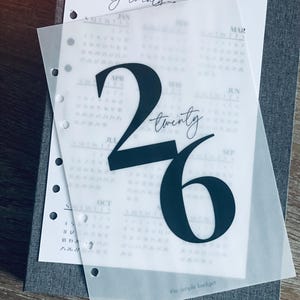 May include: A grey planner with a translucent overlay. The overlay has the number '26' in dark blue, and the text 'twenty twenty'. The calendar page underneath shows months and dates. The words 'the simple budget' are also visible.