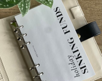 Può includere: Una pagina bianca con il testo "holiday SINKING FUNDS" in caratteri neri. La pagina è in un raccoglitore bianco con anelli argentati. Il testo "the simple budget" è visibile nell'angolo in basso a sinistra della pagina.