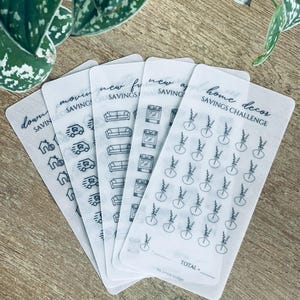 May include: Five clear sheets of stickers with black outlines of various icons. The stickers are for a savings challenge and include icons for a house, moving van, sofa, oven, and a plant. The text on the stickers reads "down payment savings", "moving savings", "new furniture savings", "new appliances savings", and "home decor savings challenge".