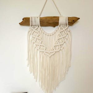 Puede incluir: Un tapiz de macramé blanco con una rama de madera natural en la parte superior. El tapiz tiene un diseño de flecos y está hecho con cordón de algodón.